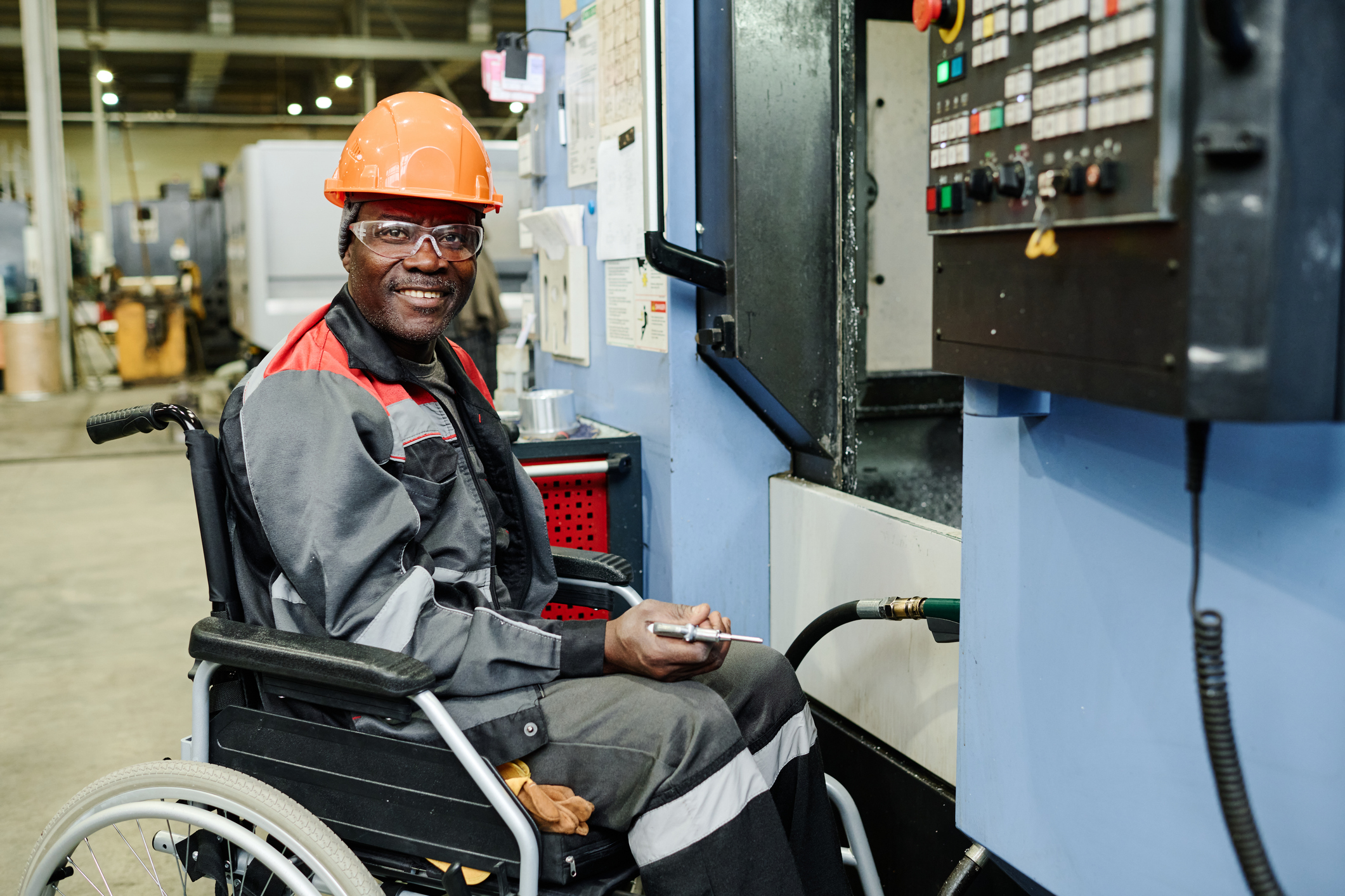 Man in wheelchair and hardhat working on electrical panel in warehouse.