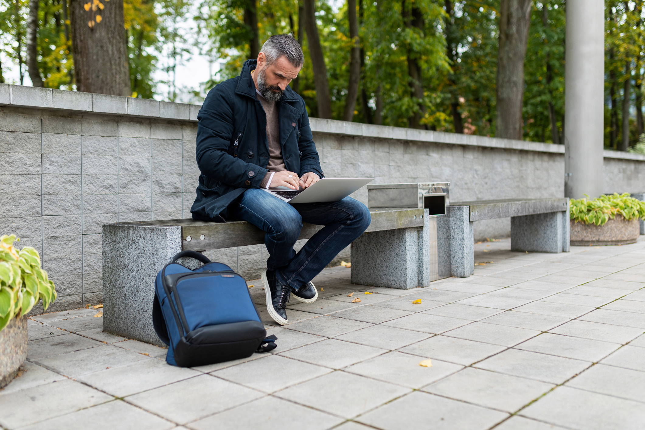 Middle-aged man sitting on bench along a city sidewalk near a park, working on a laptop.