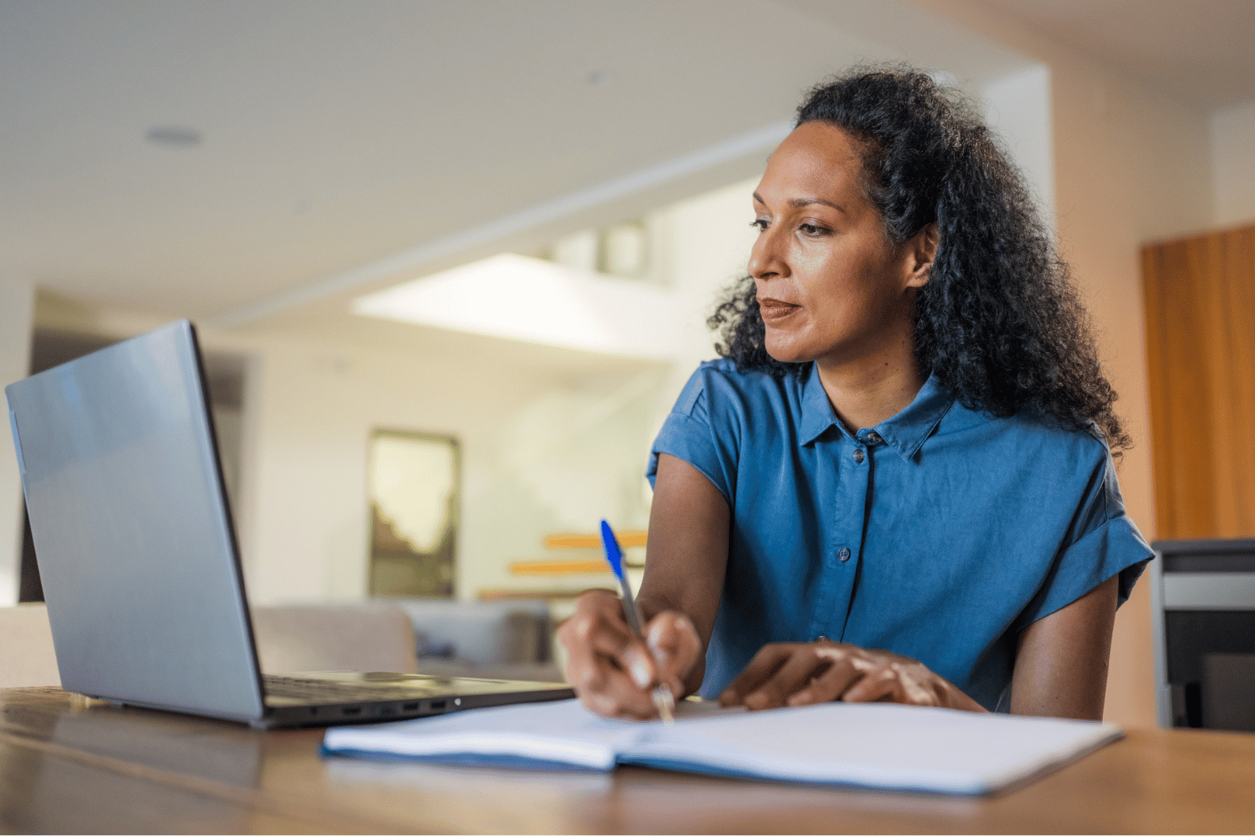 Woman sits at table in her home, writing in a notebook while looking at a laptop.
