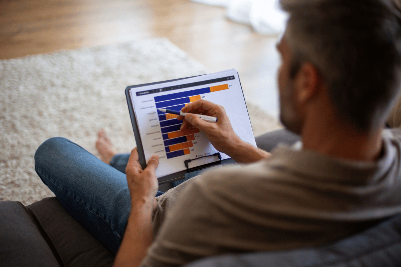 Middle-aged man on couch at home looks at a bar graph of figures on a digital pad.