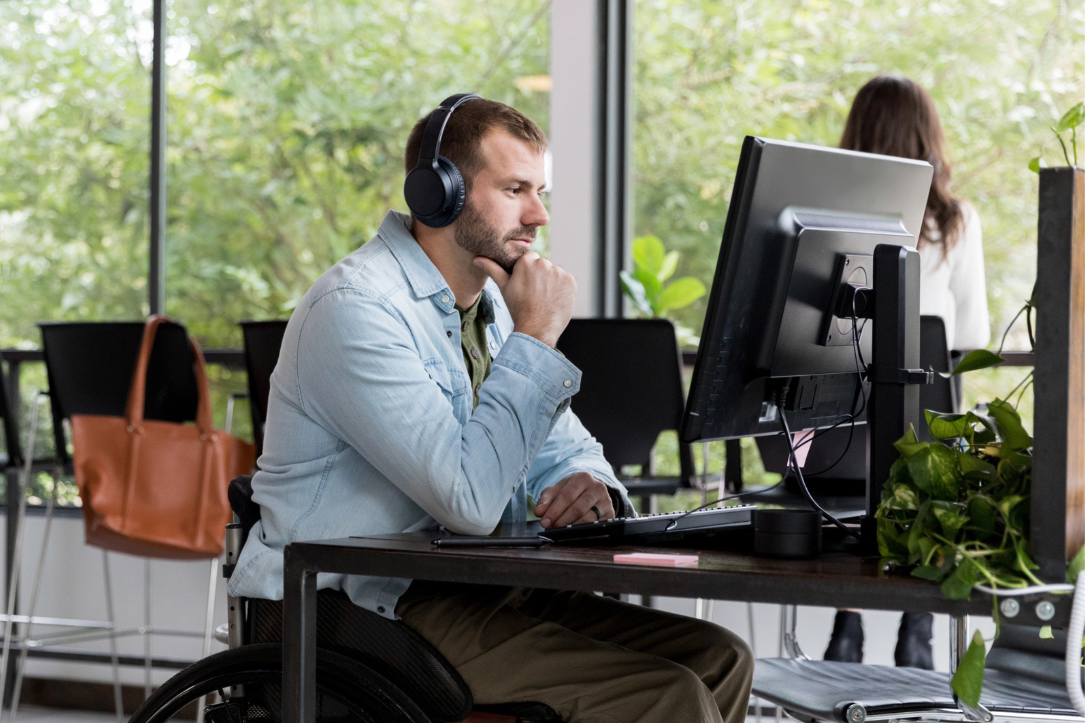 Man in his 30's working on a computer in an office, seated in wheelchair at his desk with headphones on.