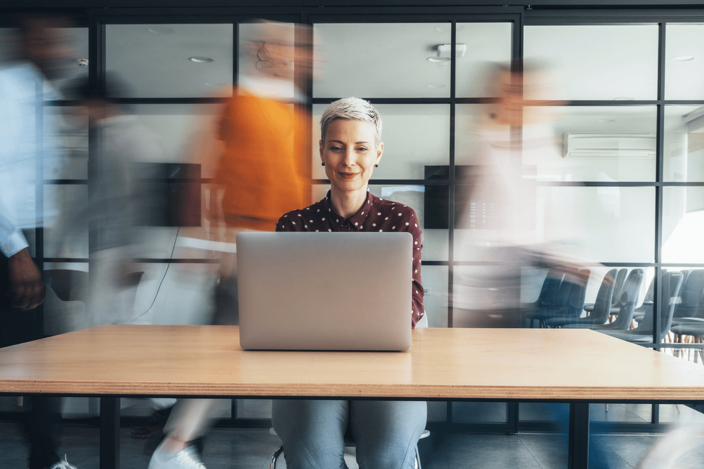 Middle-aged woman sitting at laptop in office while others rush around behind her.