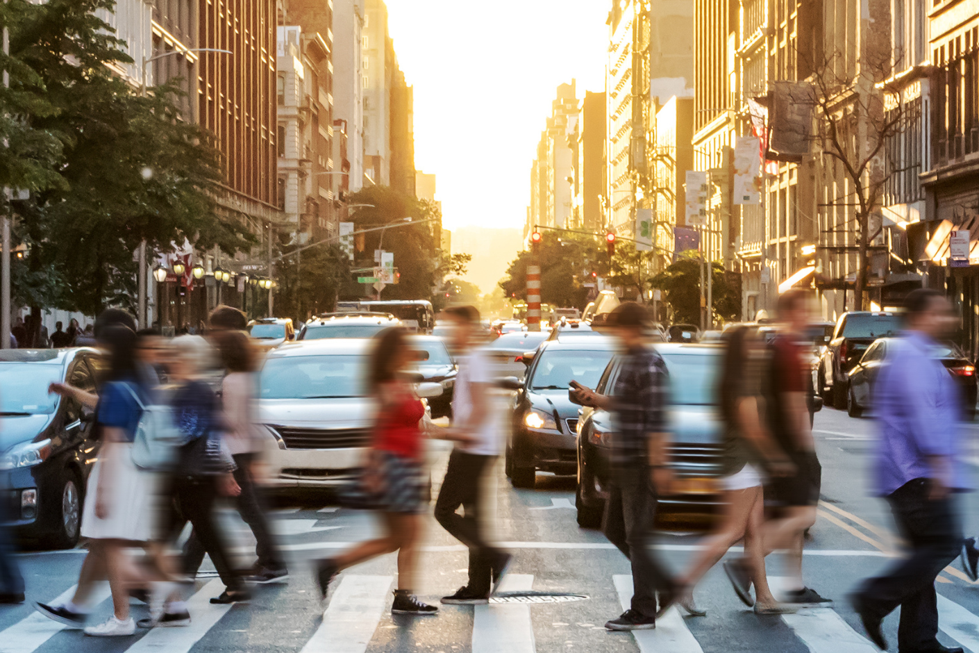 Looking down a busy city street from the middle of an intersection, with a motion blur of many people crossing left and right in the foreground.