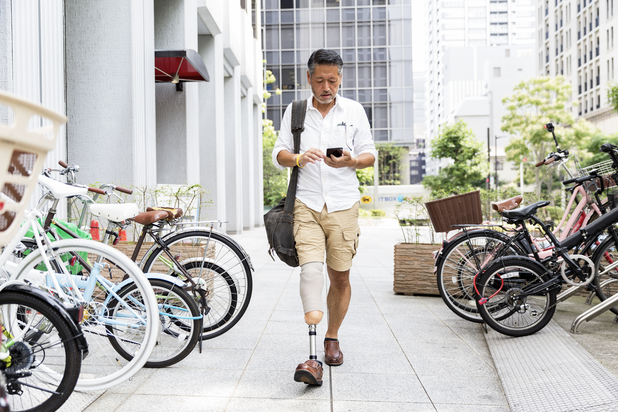 Man in business-casual dress with a prosthetic leg walks down a city sidewalk while checking his smartphone.