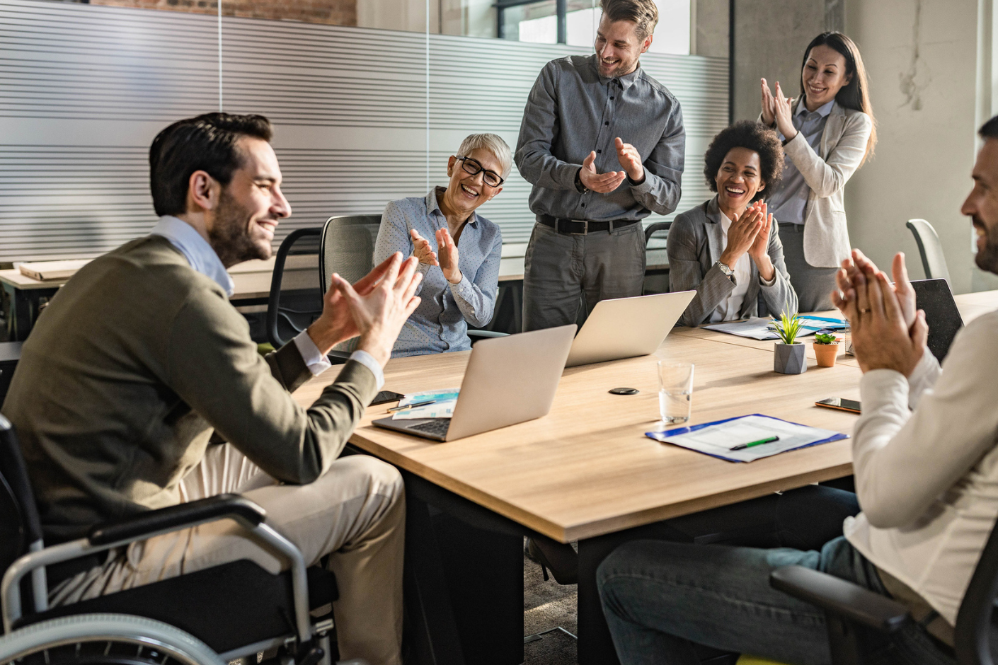 Man in wheelchair sits at the head of a conference table, being applauded by peers in a business meeting.