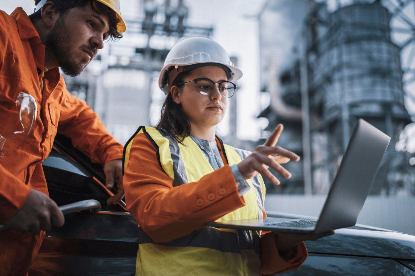 Woman in safety vest and helmet works on a laptop at an industrial job site, conferring with a workman.